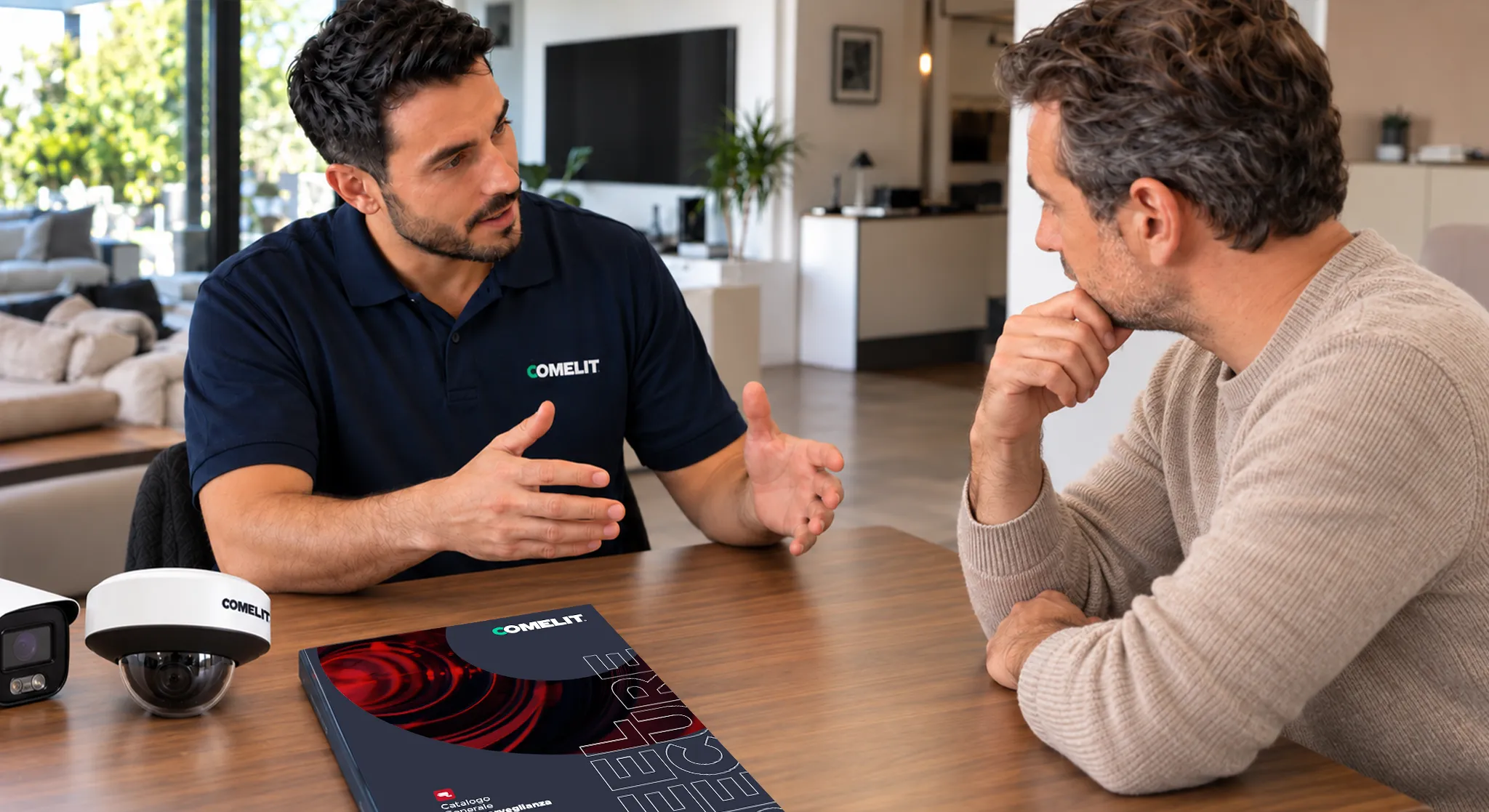 Installatore Comelit spiega a un cliente come scegliere le telecamere di videosorveglianza, con catalogo e telecamere sul tavolo